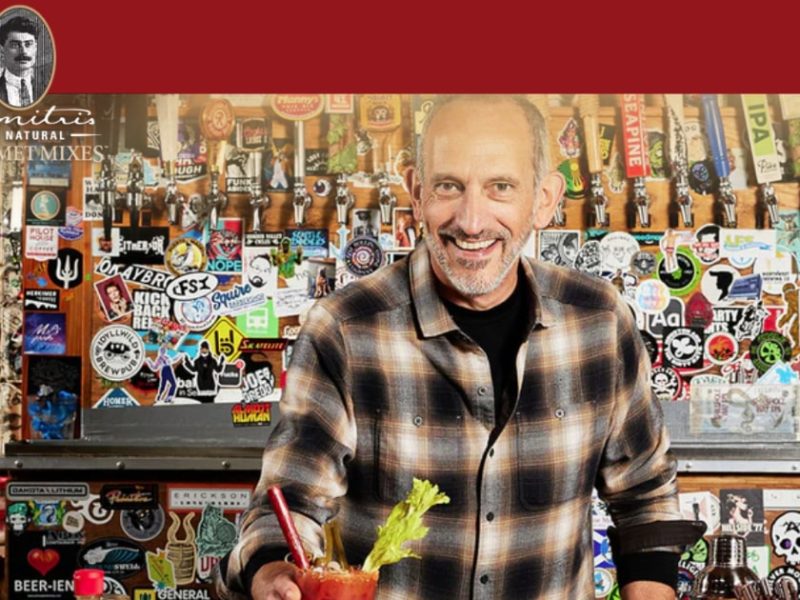Entrepreneur holding a Bloody Mary, smiling in front of a bar adorned with stickers and beer taps.
