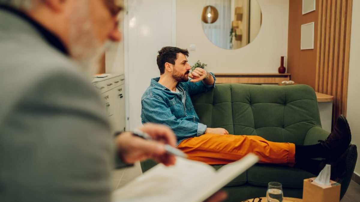 A man in a denim jacket sits on a green couch, deep in thought, while another person writes in a notebook nearby.