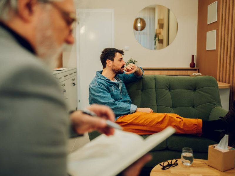 A man in a denim jacket sits on a green couch, deep in thought, while another person writes in a notebook nearby.