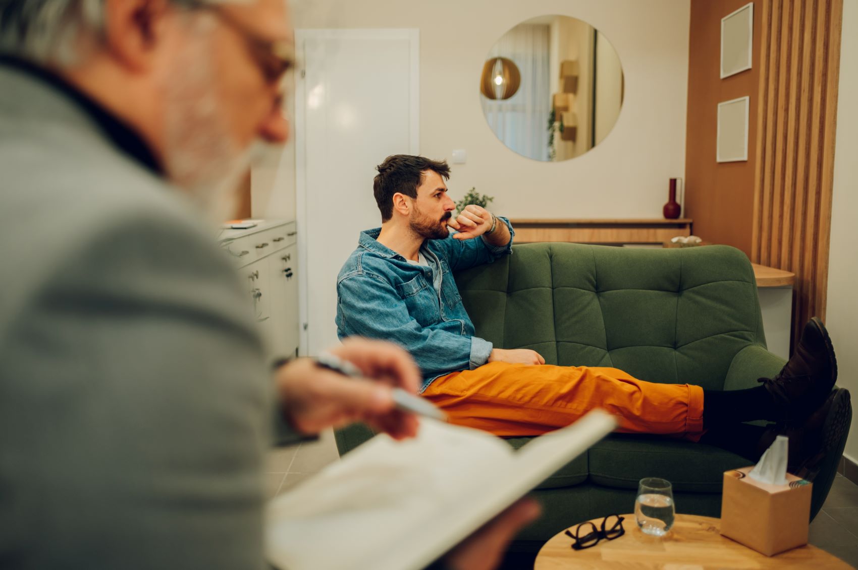 A man in a denim jacket sits on a green couch, deep in thought, while another person writes in a notebook nearby.