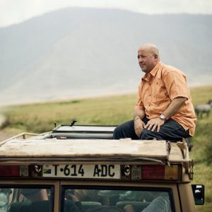 Andrew Zimmern enjoying a South African safari from the roof of a vehicle, with a landscape of grasslands and zebras in the background.