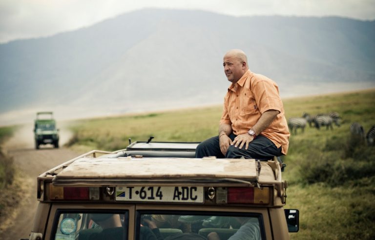 Andrew Zimmern enjoying a South African safari from the roof of a vehicle, with a landscape of grasslands and zebras in the background.