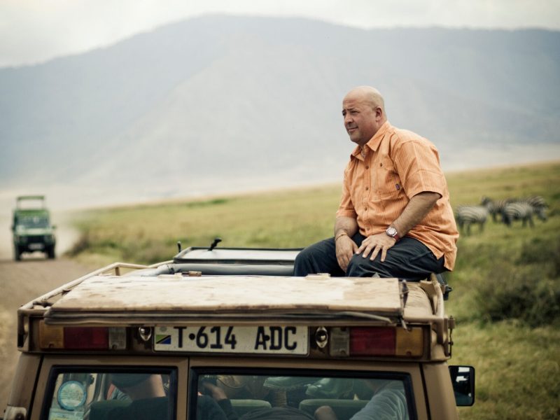 Andrew Zimmern enjoying a South African safari from the roof of a vehicle, with a landscape of grasslands and zebras in the background.