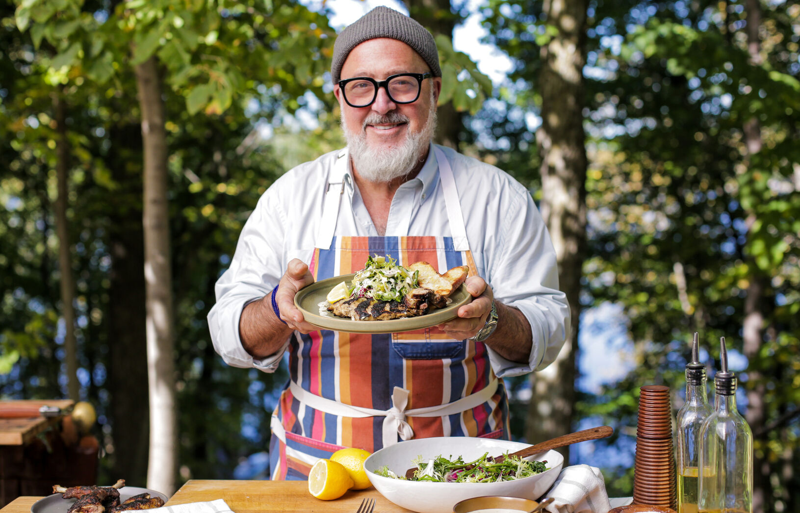 Andrew Zimmern holds a plate of grilled food, smiling in a kitchen-like outdoor setting surrounded by trees.