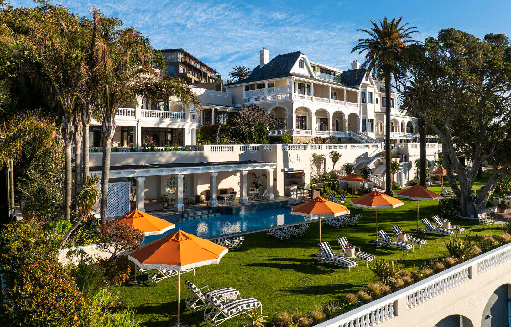 Luxurious Ellerman House hotel featuring a pool, lounge chairs, and orange umbrellas amid lush greenery in South Africa.