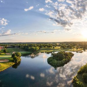 Scenic view of Lake Geneva with a golf course, trees, and a fountain under a bright sky at sunset.