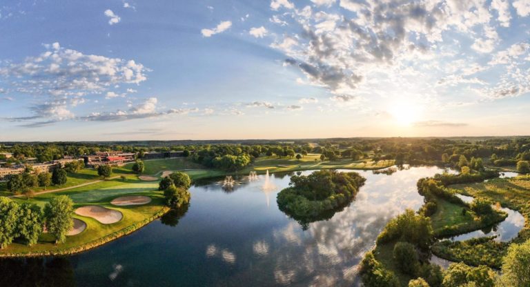 Scenic view of Lake Geneva with a golf course, trees, and a fountain under a bright sky at sunset.