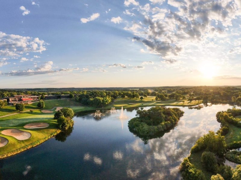Scenic view of Lake Geneva with a golf course, trees, and a fountain under a bright sky at sunset.