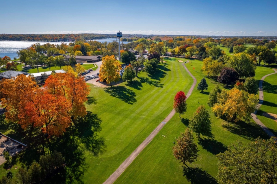 Aerial view of a vibrant golf course with colorful trees and a lake in the background, showcasing a beautiful autumn day.