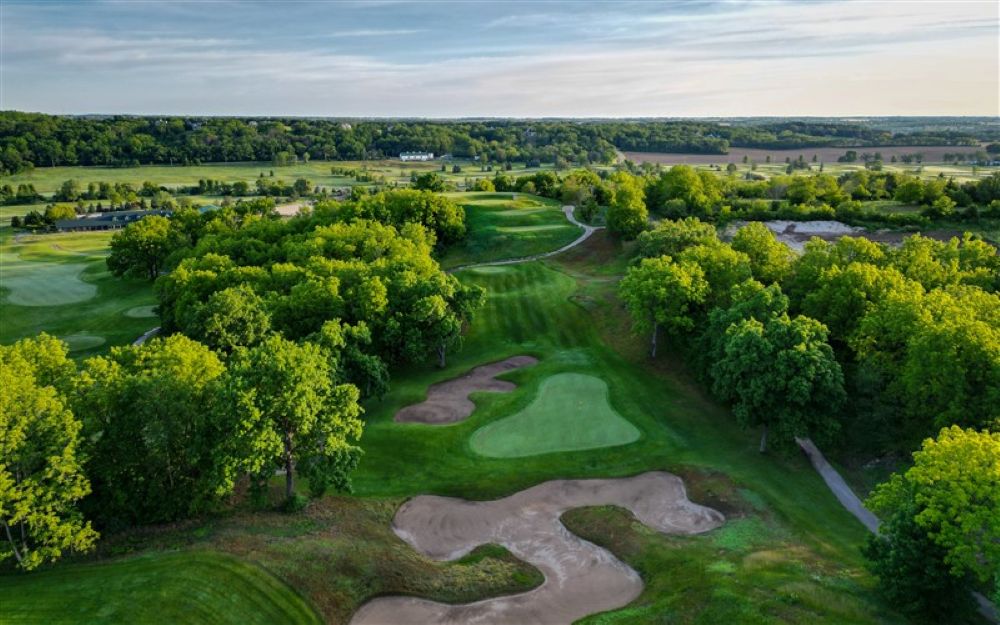 Aerial view of a lush golf course surrounded by trees, featuring manicured greens and sand traps in the foreground.