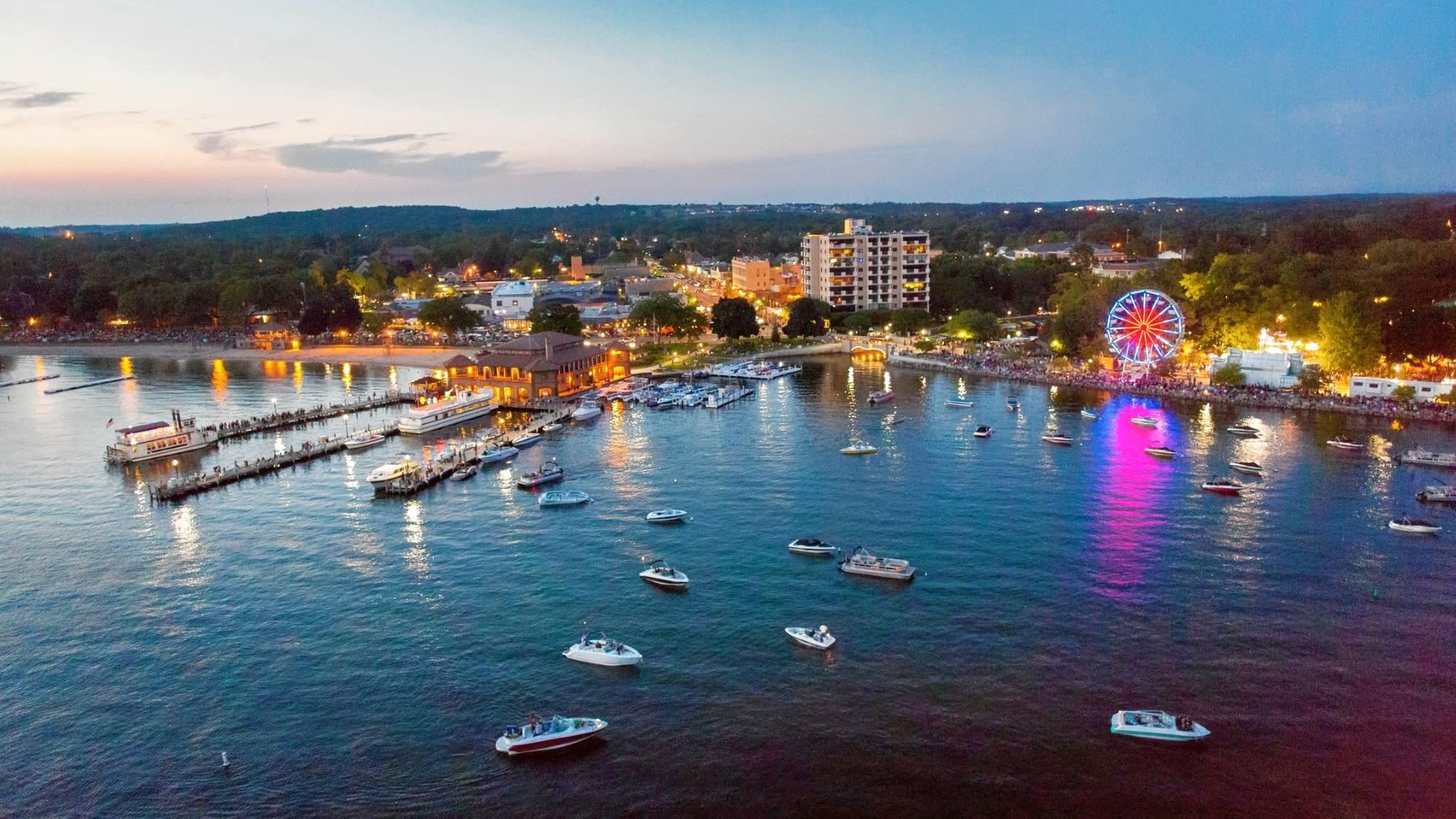 Panoramic view of Lake Geneva at dusk, featuring boats on the water and the illuminated shoreline with a ferris wheel.