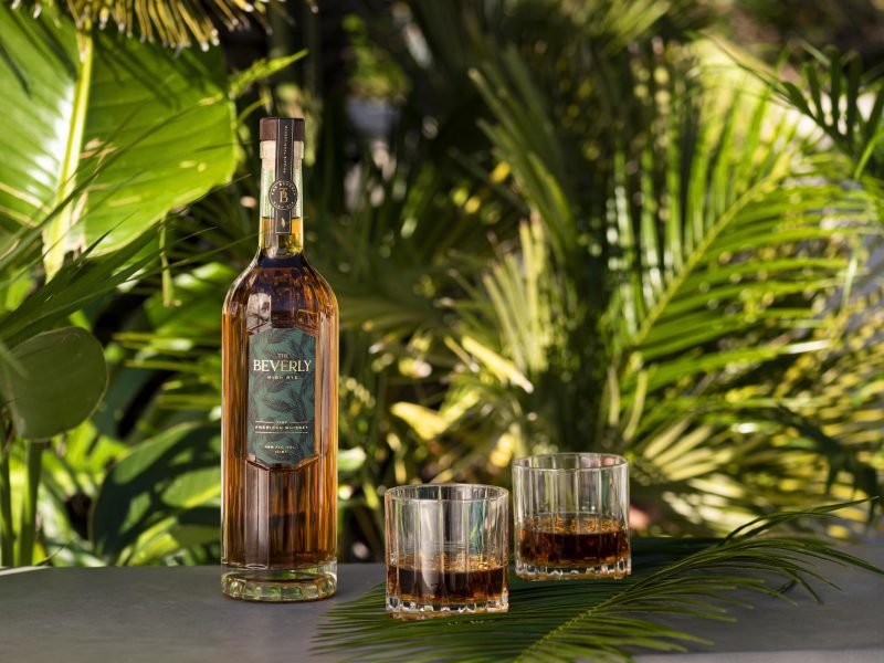 Beverly High Rye whiskey bottle with two glasses filled, set against tropical green foliage.