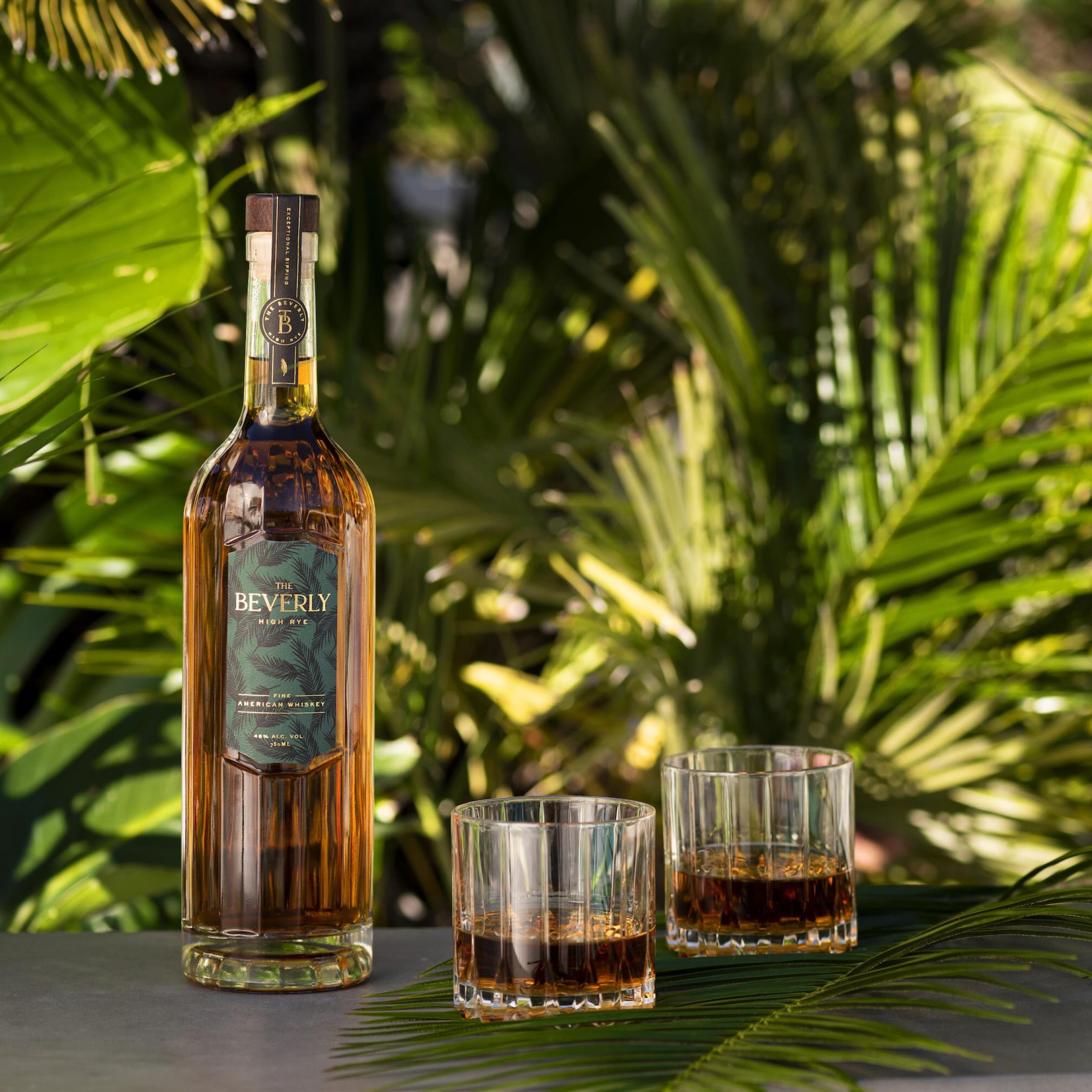 Beverly High Rye whiskey bottle with two glasses filled, set against tropical green foliage.