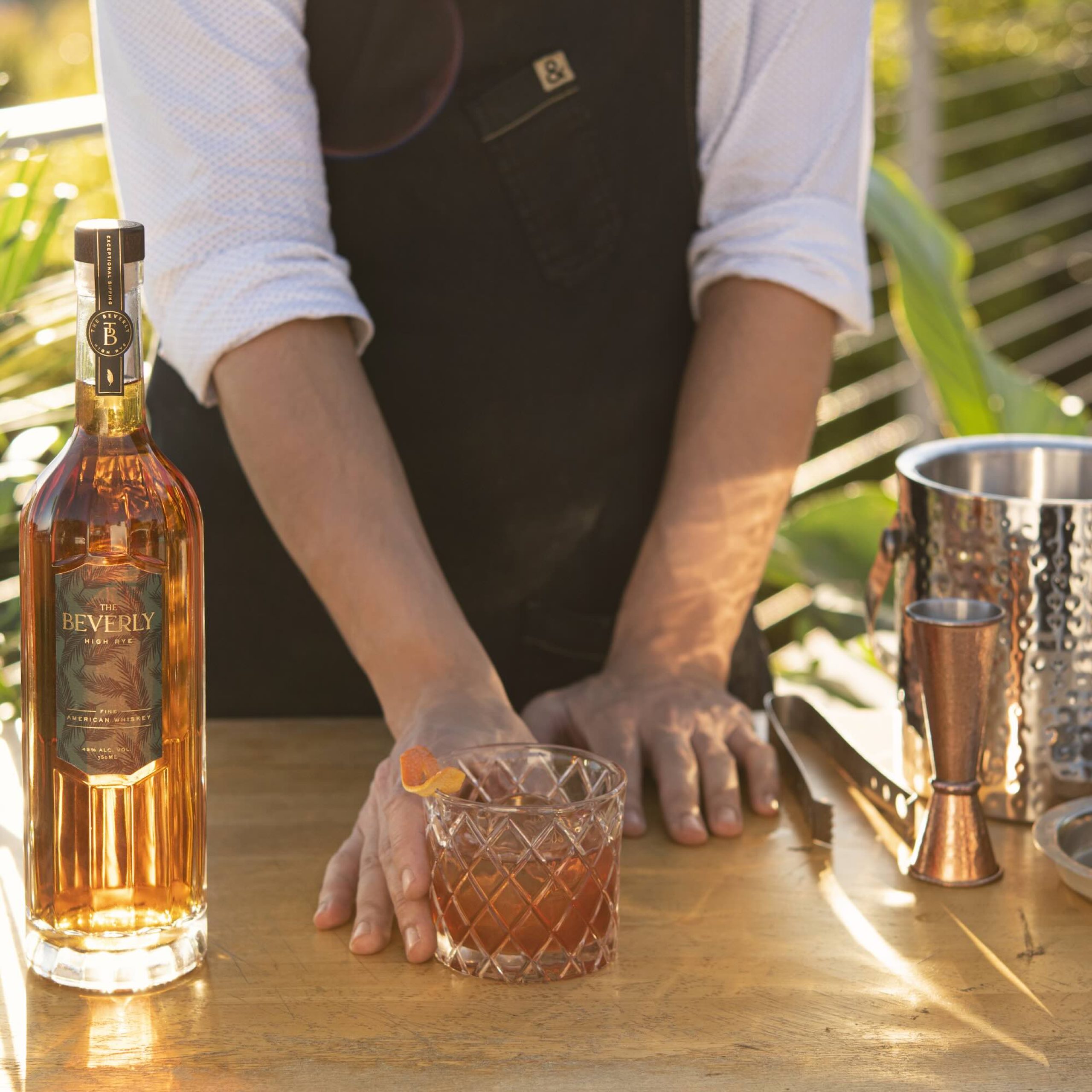 Beverly High Rye whiskey bottle beside a cocktail glass on a wooden table, surrounded by greenery and bartending tools.