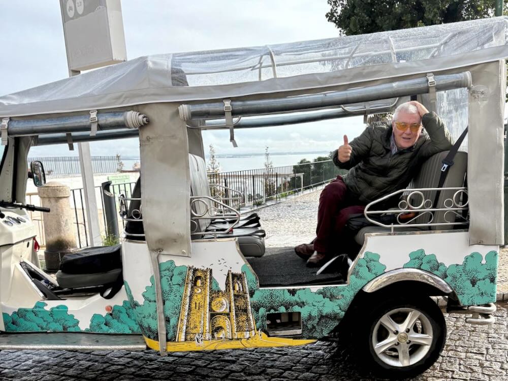 An elderly man gives a thumbs up while sitting in a colorful tuk-tuk on a scenic street in Lisbon.