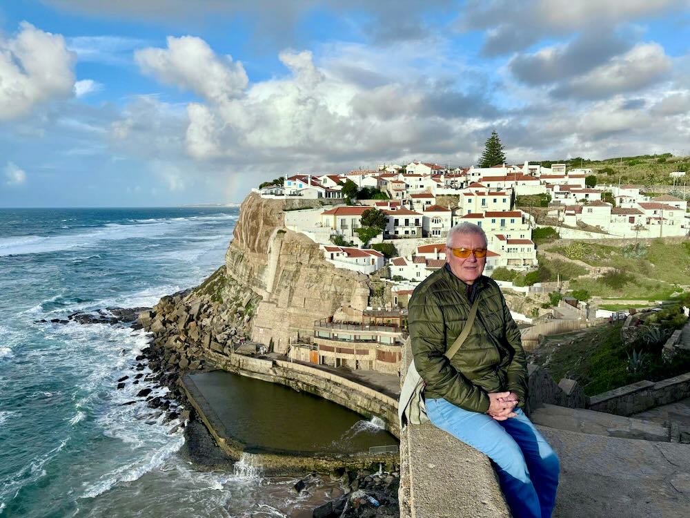 A person sitting on a wall overlooking a coastal scene with waves and white buildings in a hillside village.