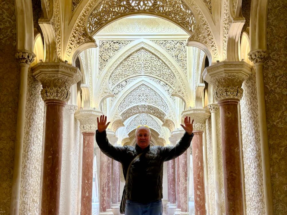 Man standing with hands raised in an ornate corridor featuring intricate arches and decorative columns.