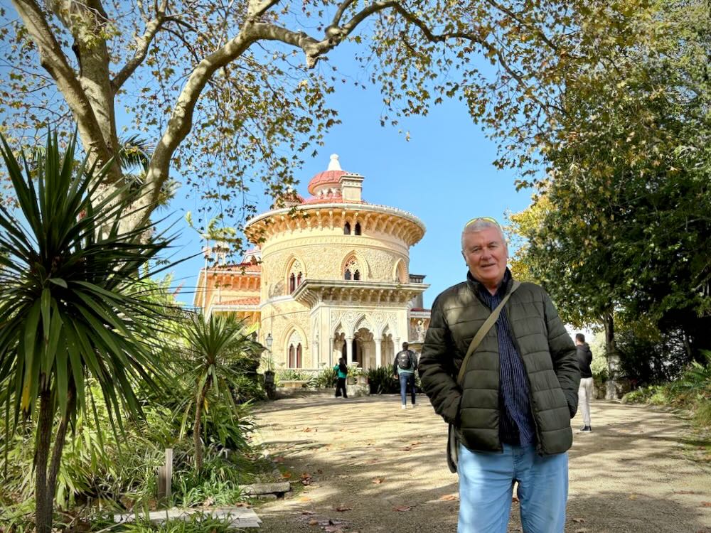 A man stands in front of a colorful, ornate building surrounded by lush greenery on a sunny day in Lisbon.