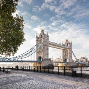 Tower Bridge spans the River Thames, surrounded by trees and a riverside walkway under a partly cloudy sky.