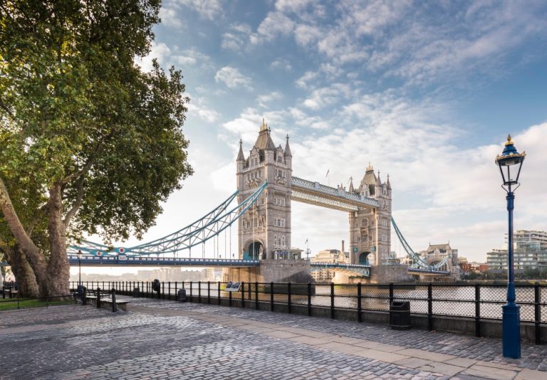 Tower Bridge spans the River Thames, surrounded by trees and a riverside walkway under a partly cloudy sky.