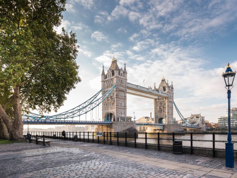 Tower Bridge spans the River Thames, surrounded by trees and a riverside walkway under a partly cloudy sky.