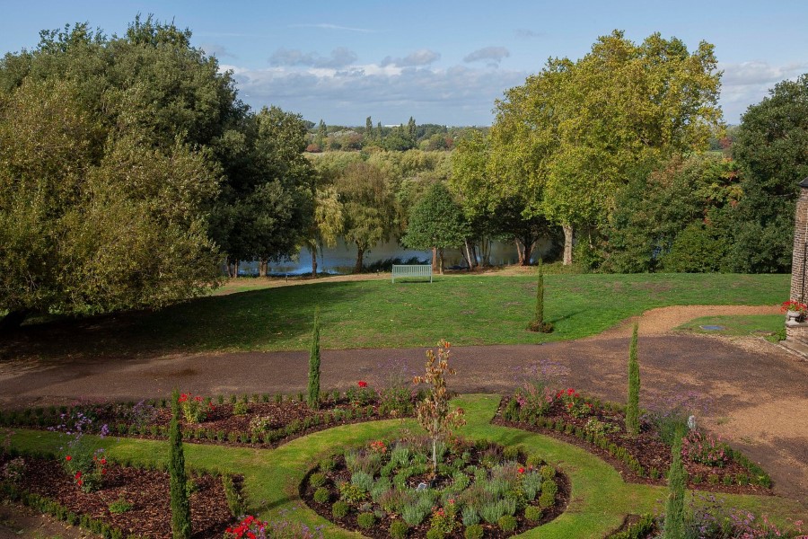 A beautiful garden with circular flower beds, trees, and a serene view of a river at Oatlands Park Hotel.