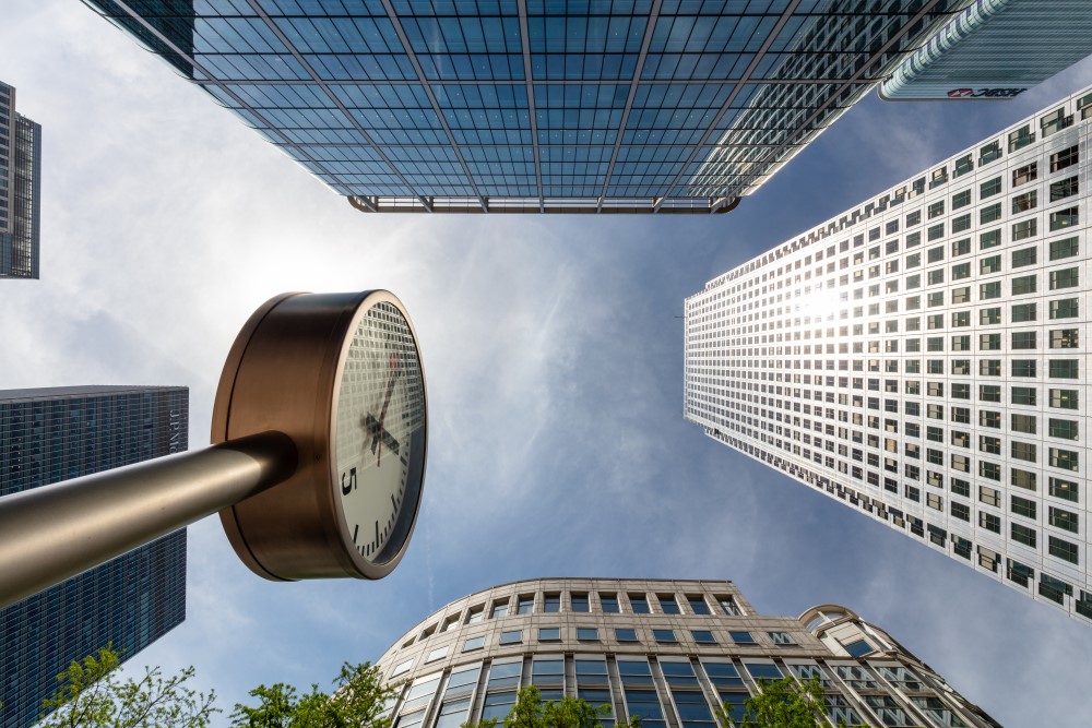 View of tall buildings and a clock from below, capturing the architectural elegance of Canary Wharf against a cloudy sky.