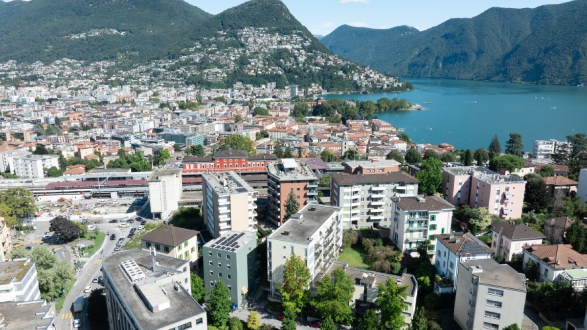 Aerial view of Lugano showcasing residential areas, mountains, and the lake with clear blue skies.