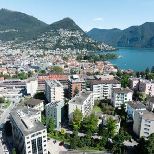 Aerial view of Lugano showcasing residential areas, mountains, and the lake with clear blue skies.