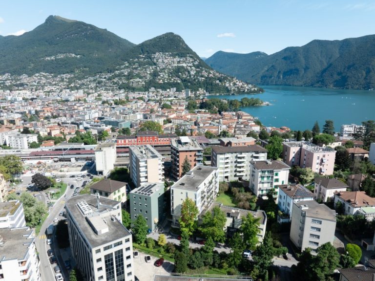 Aerial view of Lugano showcasing residential areas, mountains, and the lake with clear blue skies.