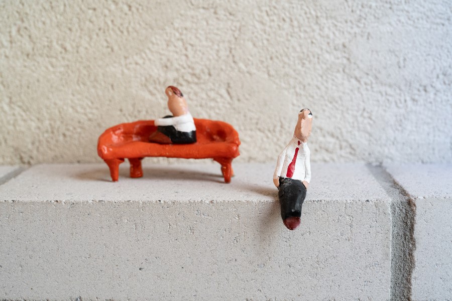 Two figurines, one seated on a stylized orange couch and the other sitting on a concrete block, create a whimsical scene.