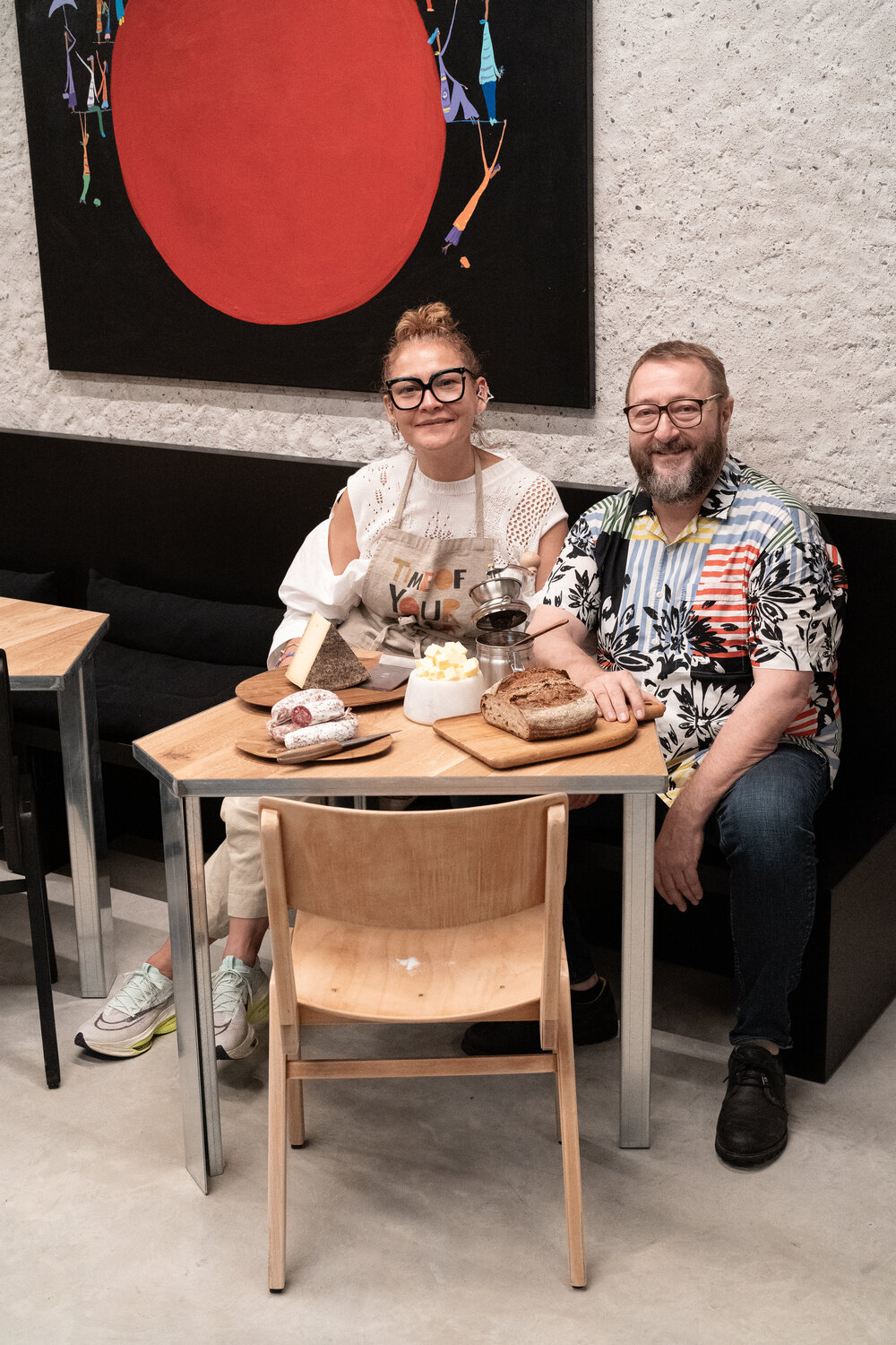 Two people sitting at a wooden table with food, smiling in a stylish cafe with modern decor and an abstract painting behind them.