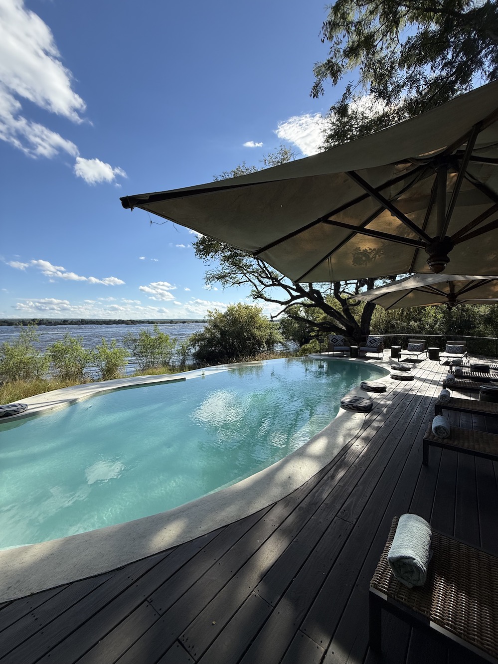 A tranquil poolside view overlooking the river, framed by trees and a bright blue sky with scattered clouds.