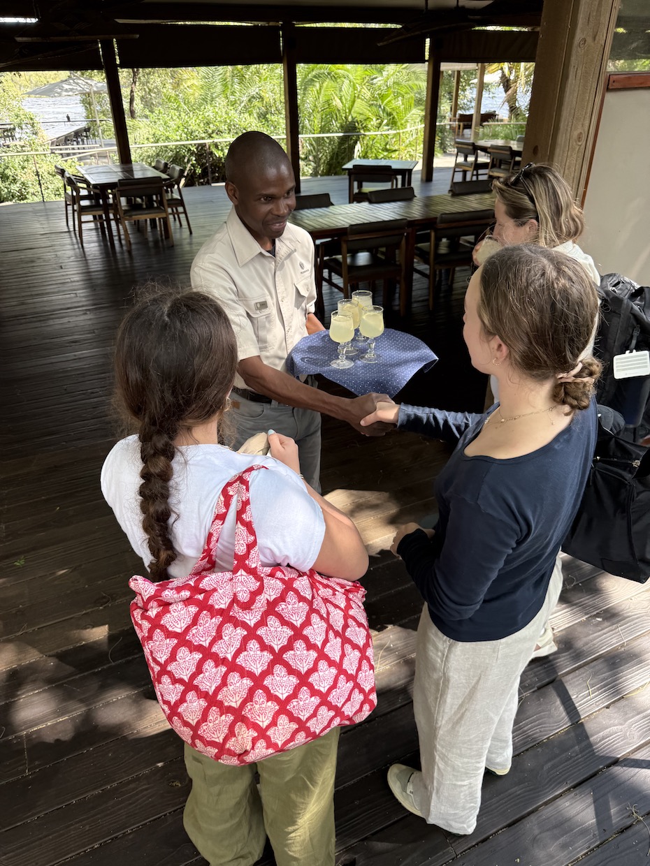 A guide serves drinks to travelers at a lodge in Zambia, with tropical greenery visible in the background.