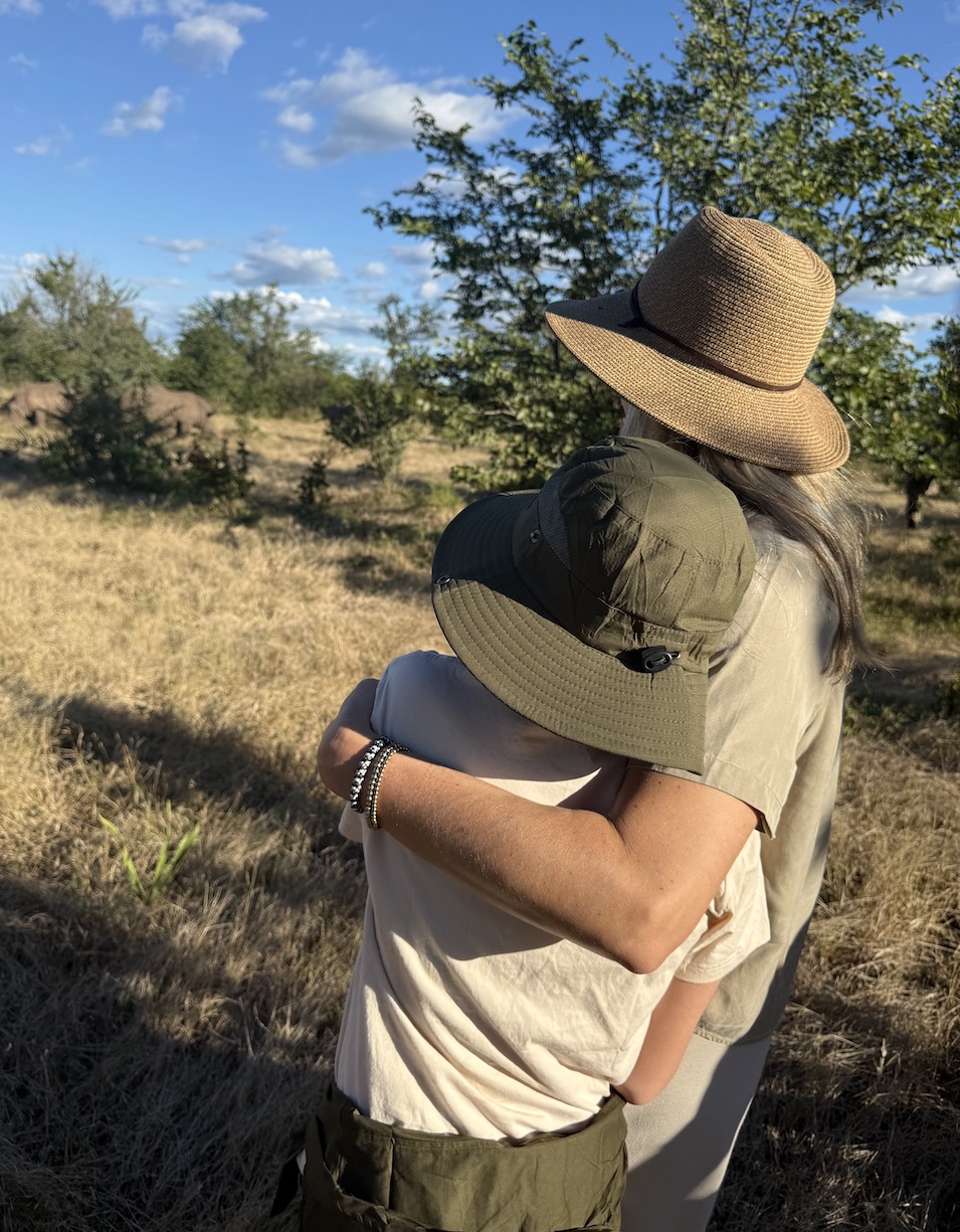 A woman and child embrace while watching wildlife in a grassy area of Zambia under a clear blue sky.