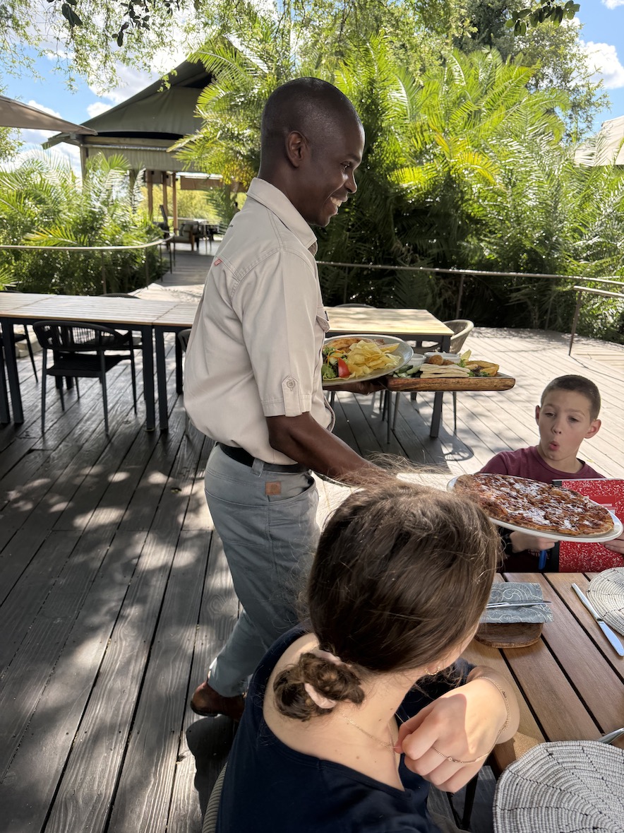 A server presents a plate of food to guests seated at a dining table surrounded by lush greenery in Zambia.
