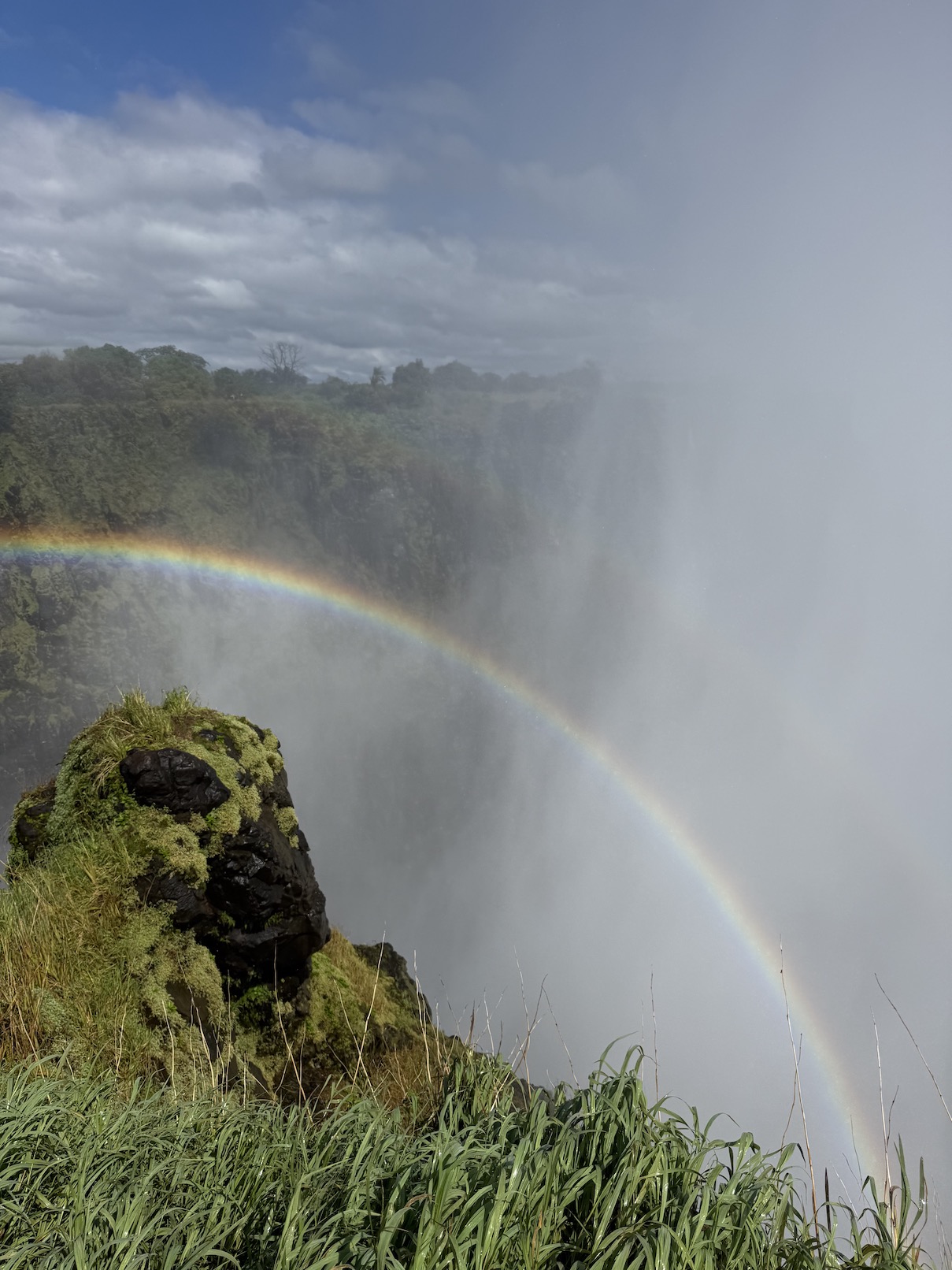 A vibrant rainbow arches over a rocky ledge amidst mist, set against a cloudy sky near a waterfall in Zambia.