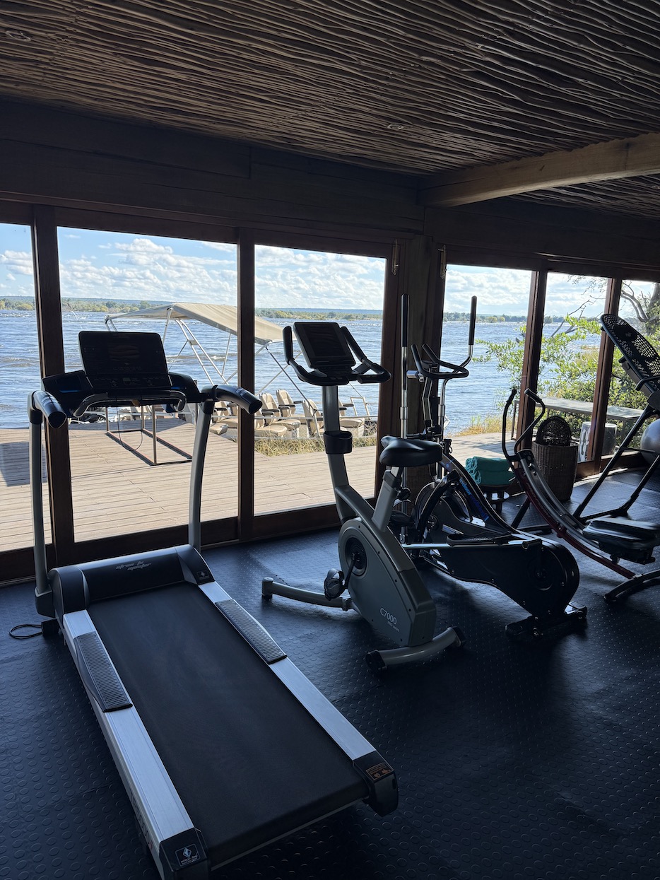 Fitness equipment including a treadmill and stationary bike overlooks a scenic view of water and sky in Zambia.