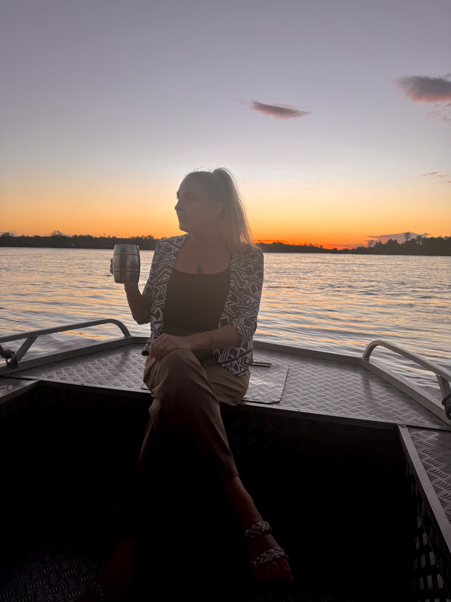 A woman sits on a boat at sunset, holding a stainless steel mug, with a tranquil river and silhouetted trees in the background.