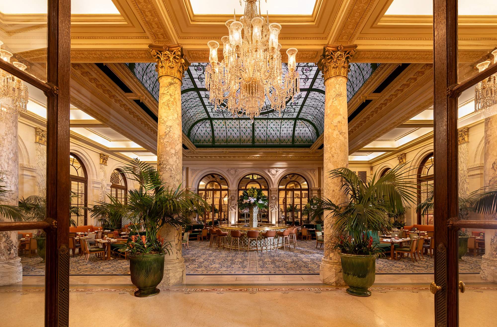 Elegant interior of the Palm Court at The Plaza, featuring chandeliers, marble columns, and lush greenery.