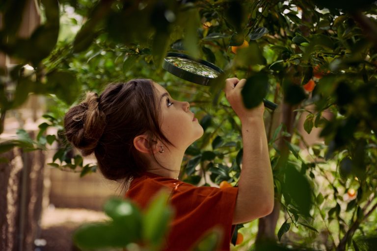 A child examines a plant closely with a magnifying glass in a lush, green environment.