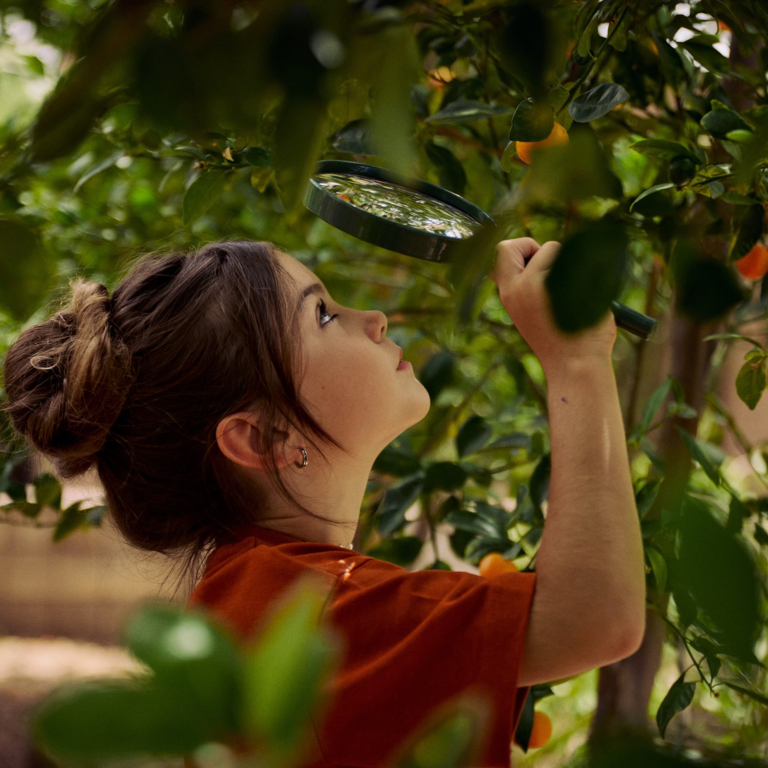 A child examines a plant closely with a magnifying glass in a lush, green environment.