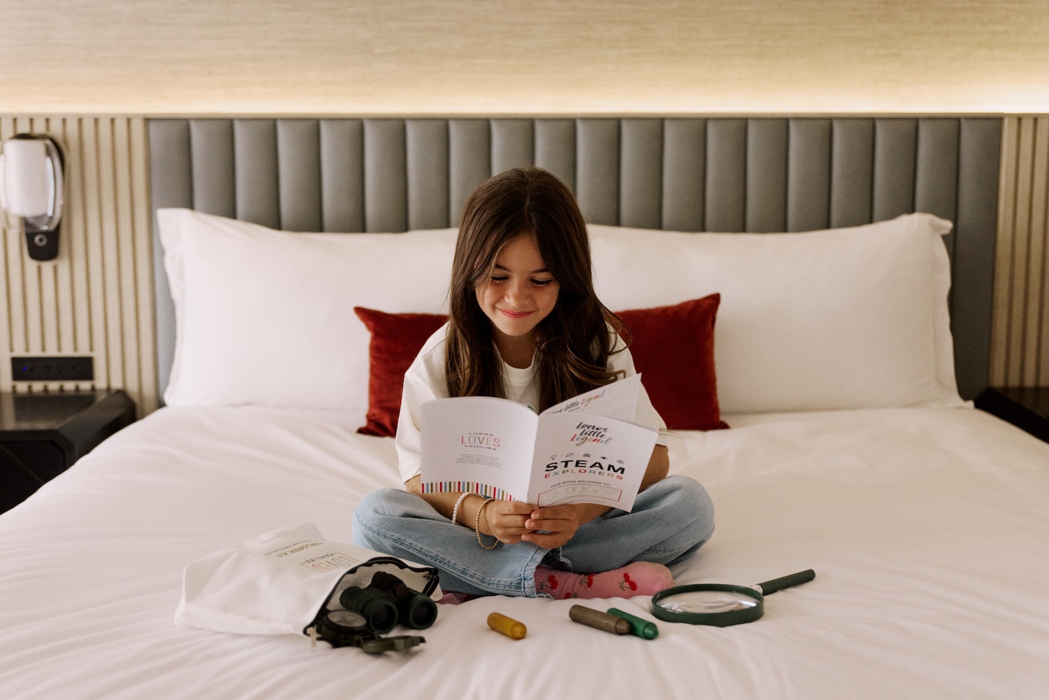 A child reads a brochure about STEAM Family Adventures while sitting on a hotel bed surrounded by exploration tools.