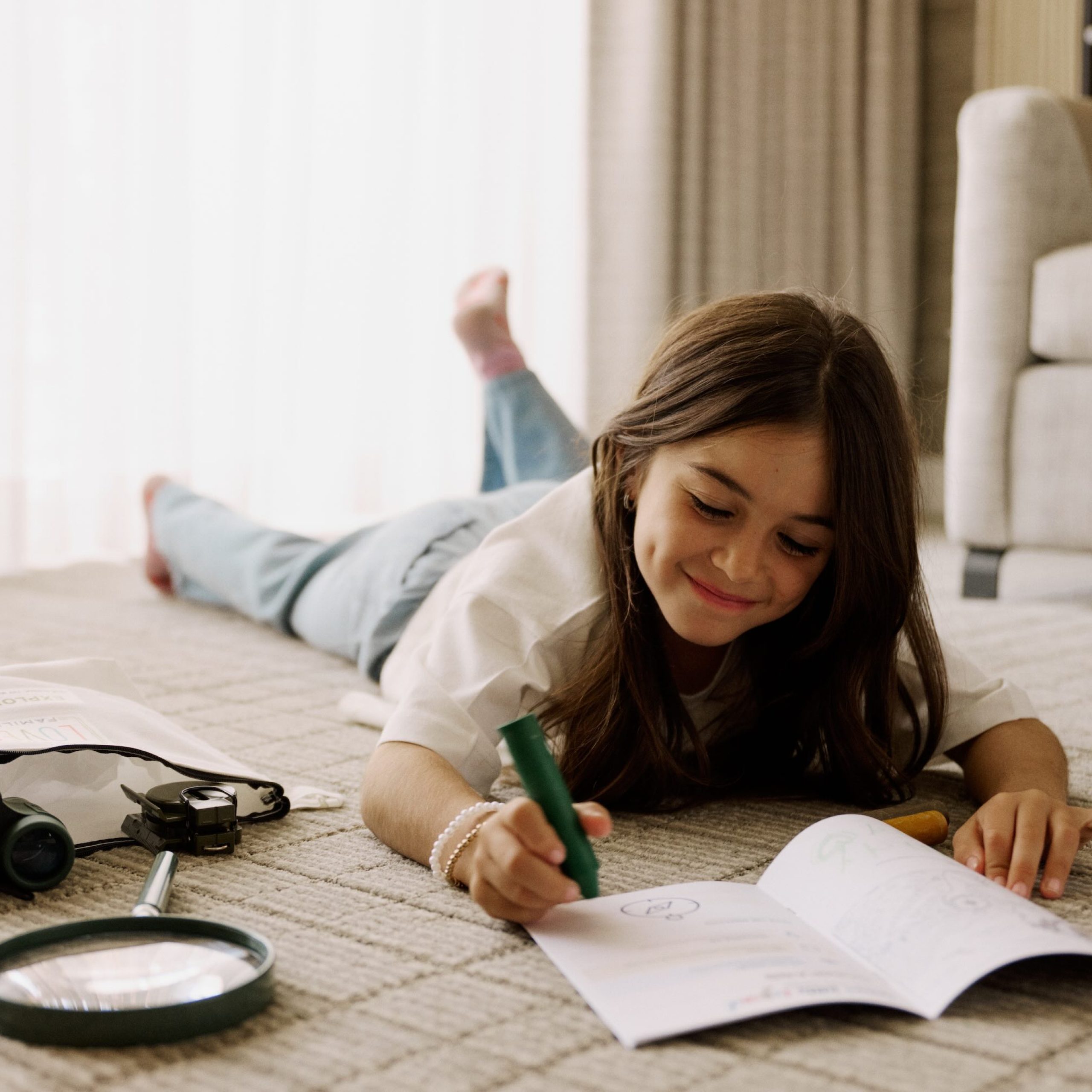 A child lying on the carpet uses a green marker to draw in a notebook near exploration tools like binoculars and a magnifying glass.