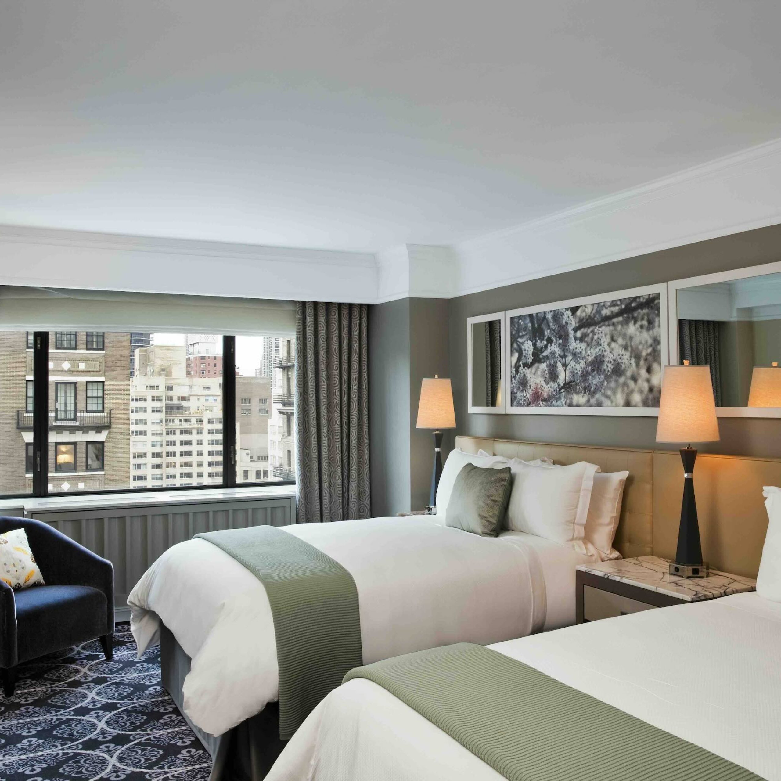 Two twin beds with green accents, a bedside lamp, and a view of the city through a large window in a modern hotel room.
