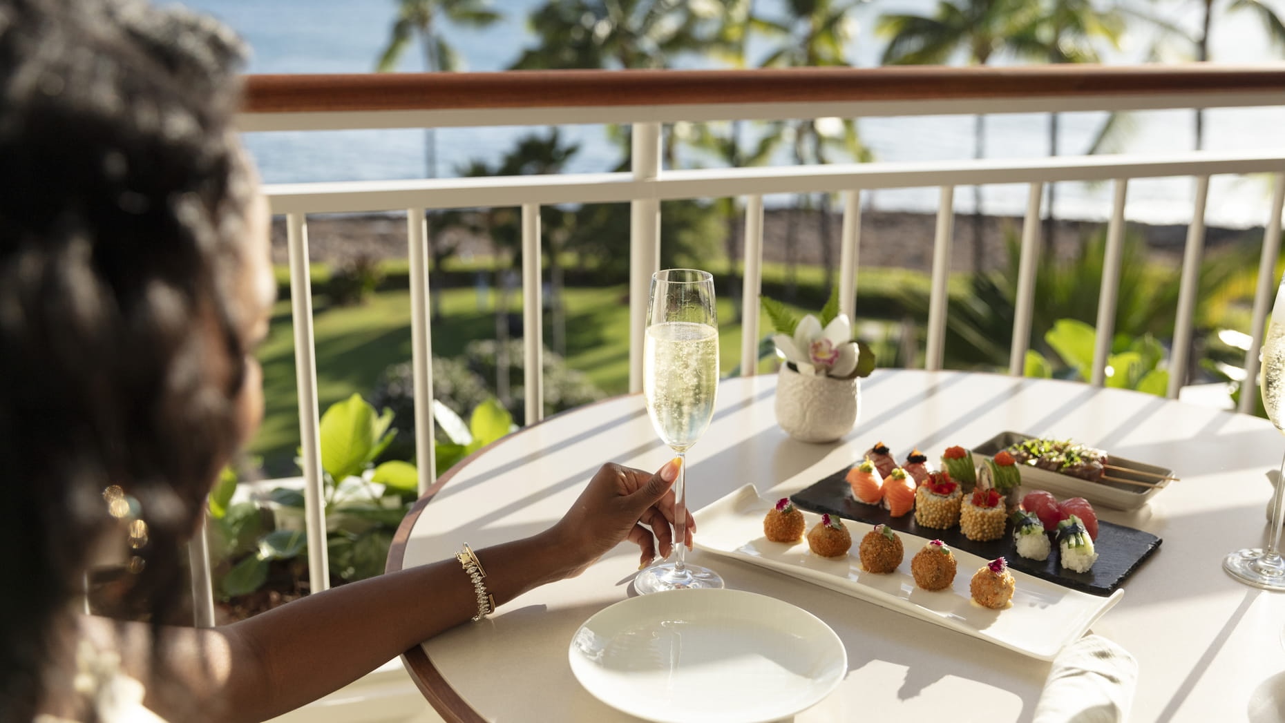 A woman enjoys drinks and appetizers on a balcony with a view of the ocean and palm trees at Four Seasons Oʻahu.