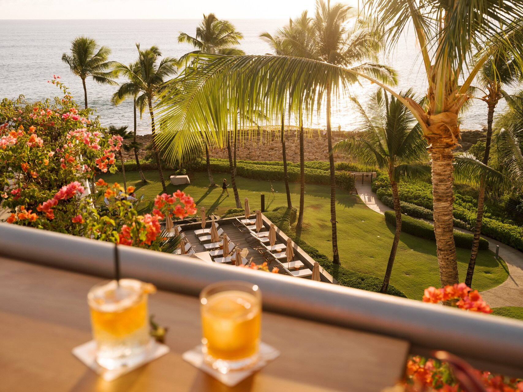 Tropical view from the Four Seasons O'ahu, featuring palm trees, vibrant flowers, and cocktails on a table at sunset.