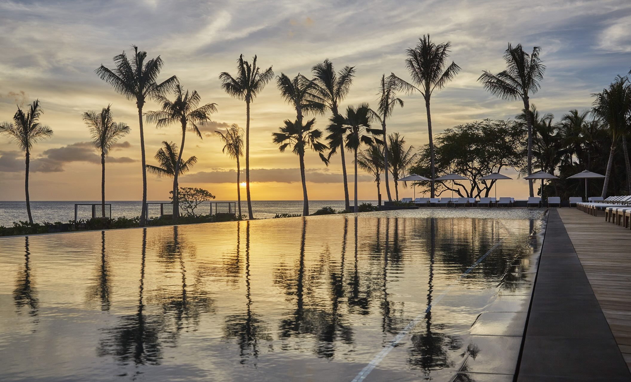 Sunset over a serene pool at Four Seasons O'ahu, with palm trees reflecting in the water and lounge chairs lining the deck.