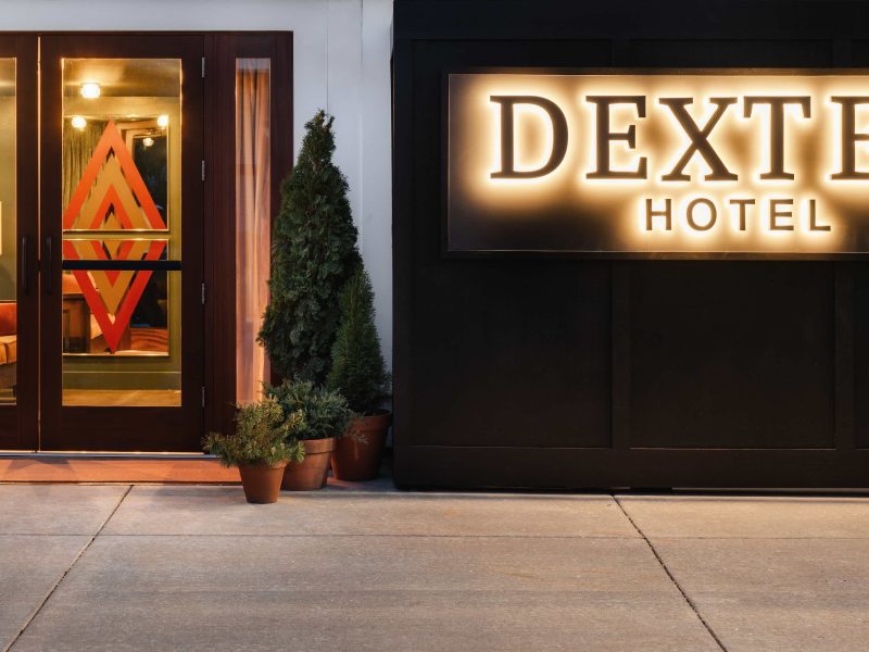 The entrance of Dexter Hotel features a stylish design with a illuminated sign and potted greenery along the walkway.