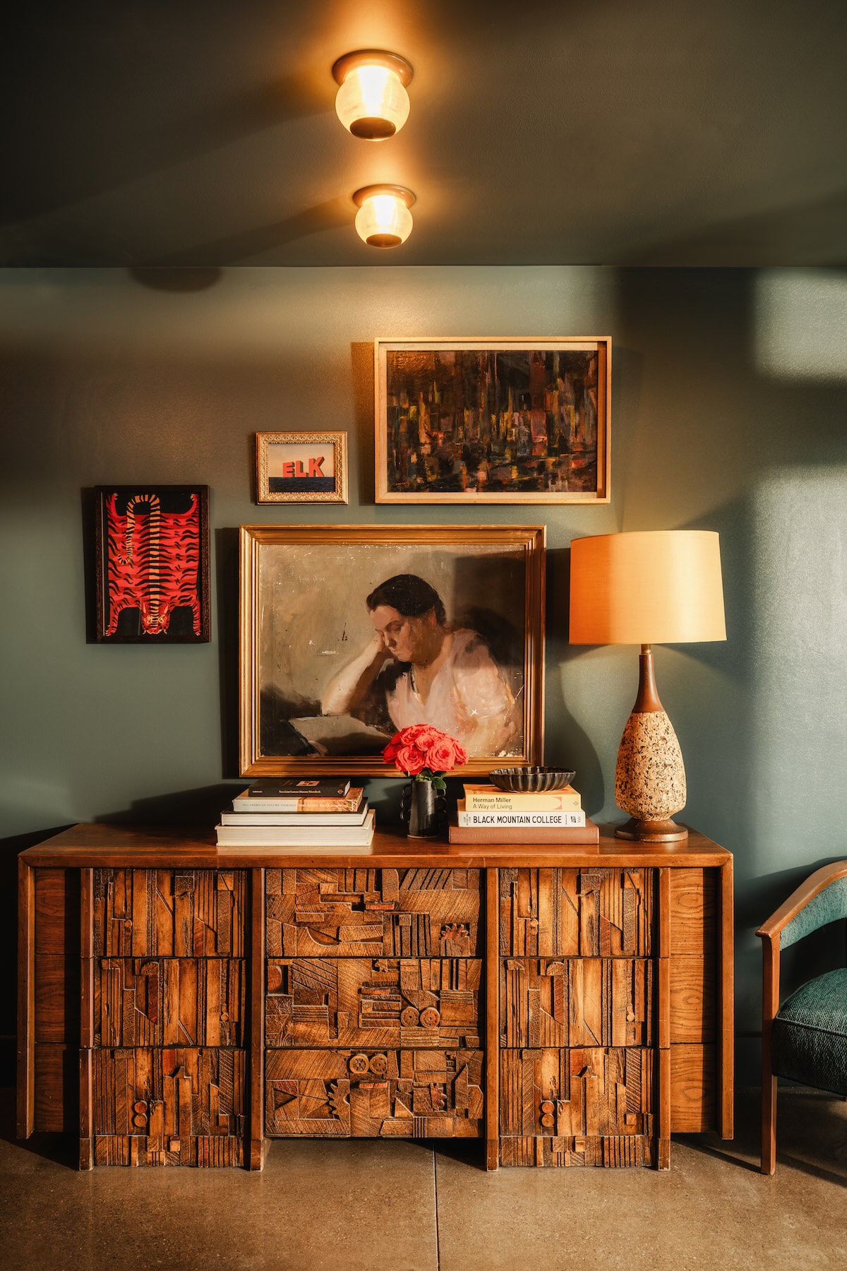A wooden sideboard adorned with books, decorative items, and a framed painting, complemented by a lamp and fresh flowers.